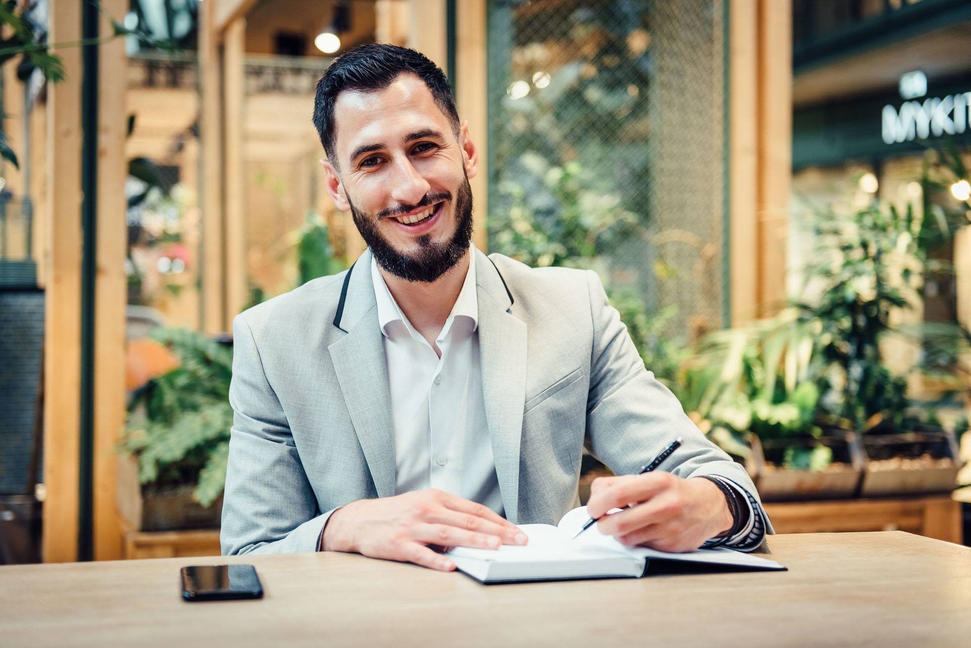 junger Mann mit Bart sitzt in einem Cafe und schreibt in sein Notizbuch