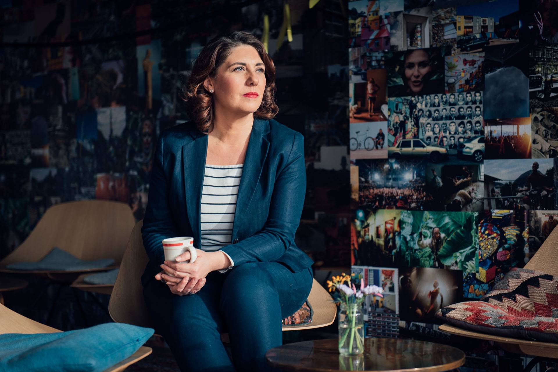 Eine Frau mit einer Kaffeetasse in der Hand sitzt vor einer bunten Wand mit vielen Bildern darauf