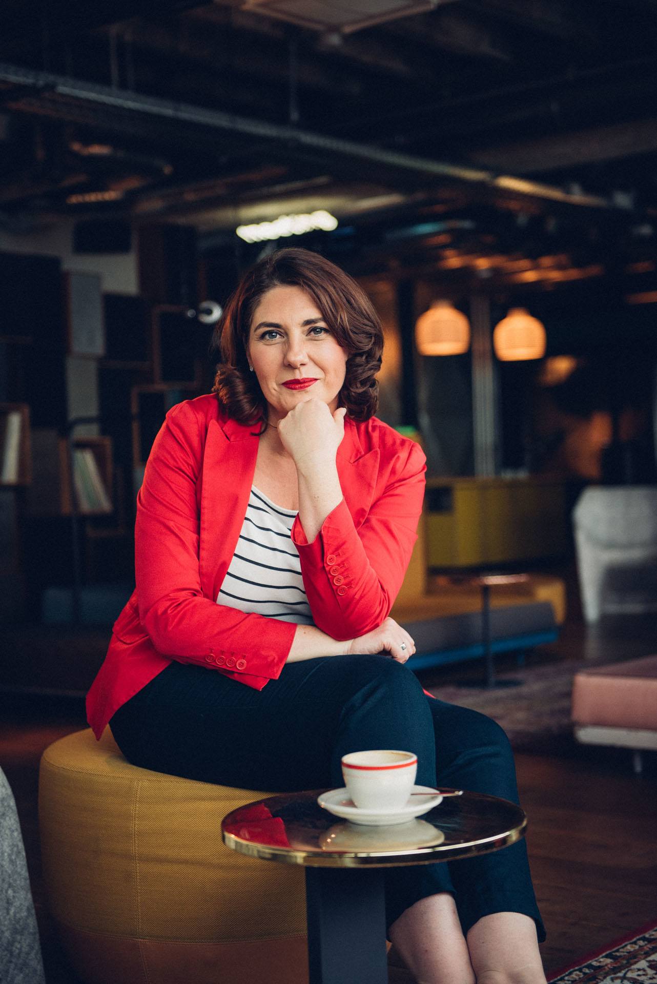 Ein weiblicher Coach mit rotem Blazer sitzt in einem Cafe und lächelt in die Kamera