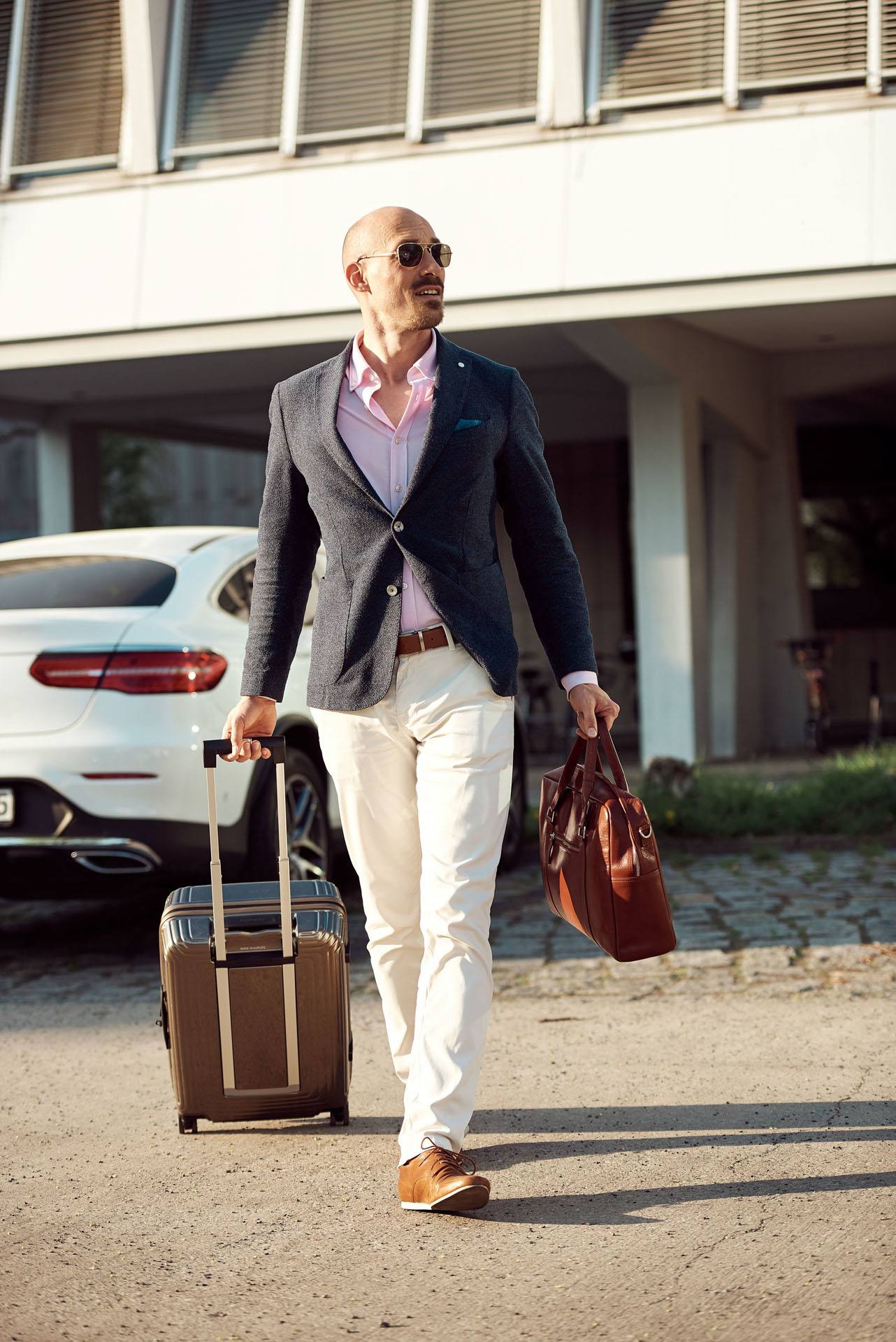 junger Mann mit Sonnenbrille, Anzug und Rollkoffer vor teurem weißen Auto